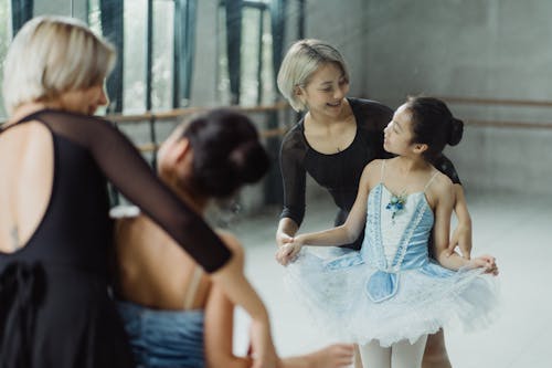 Cheerful Asian teacher standing near mirror behind back of content girl in tutu and looking at each other in ballet class