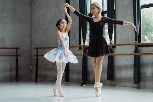 Full body of girl in tutu rehearsing dance while holding hand of teacher in pointe shoes in ballet studio during rehearsal