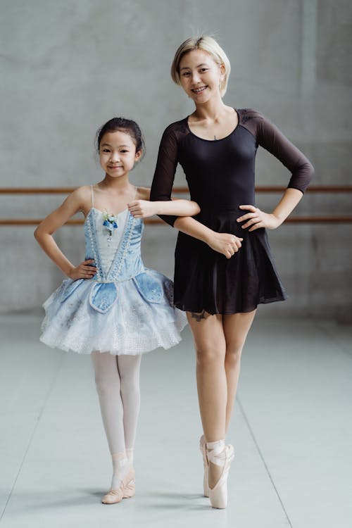 Full body of Asian teacher in pointe shoes standing with girl in tutu standing in ballet studio with barre and looking at camera