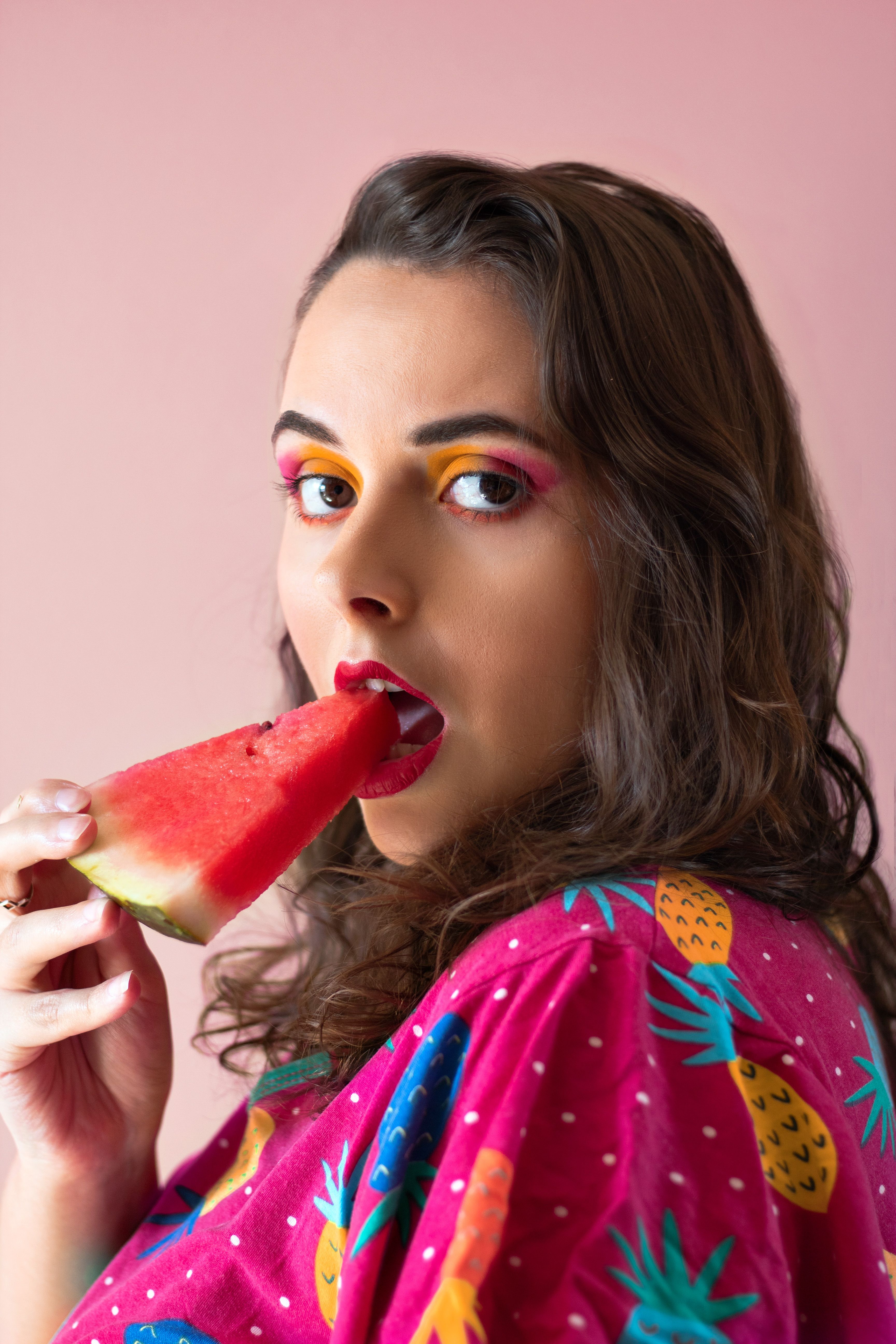 Stylish woman with vibrant makeup enjoys watermelon against a pink background.