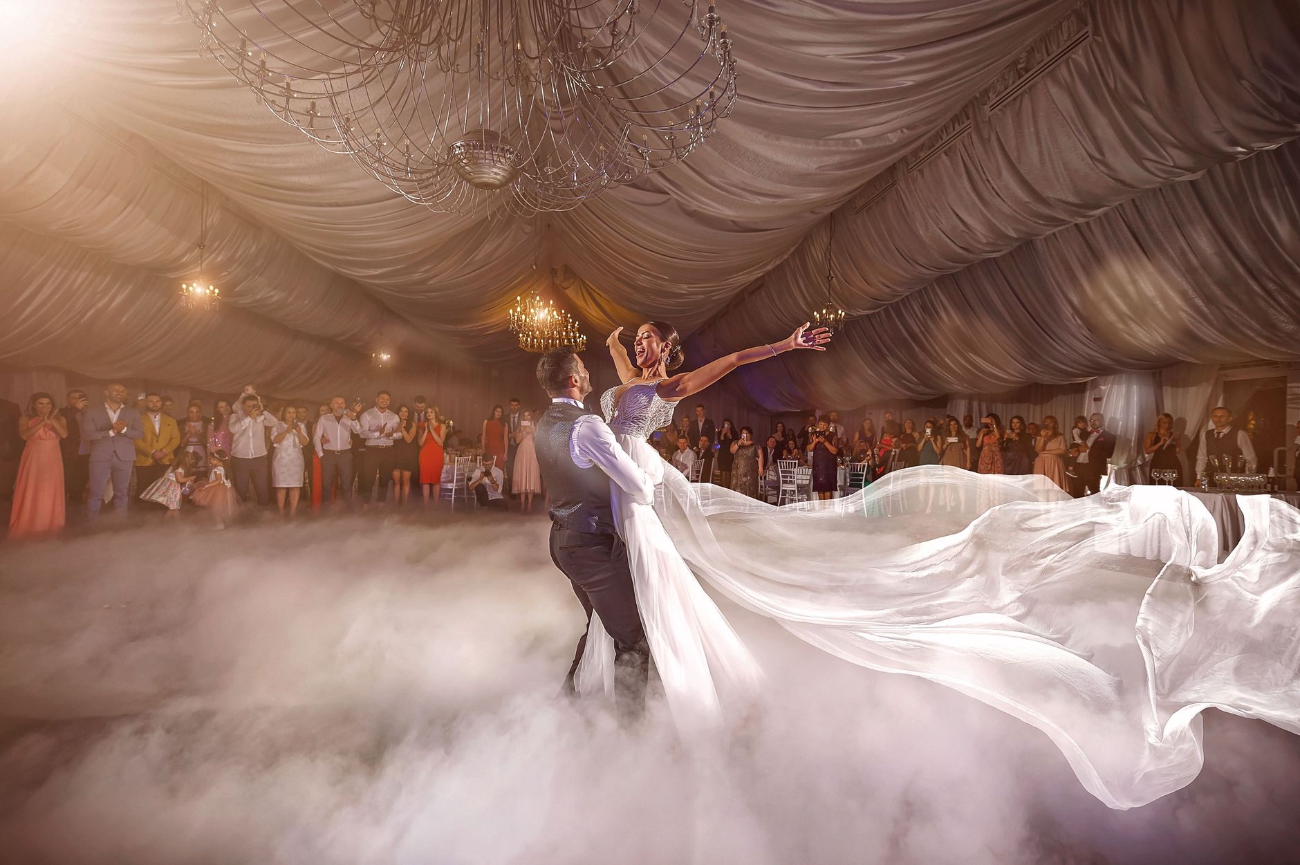 Bride and groom dance in a luxurious indoor wedding venue, surrounded by guests and ambiance.