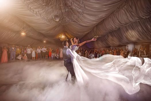 Bride and groom dance in a luxurious indoor wedding venue, surrounded by guests and ambiance.