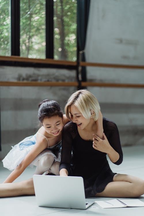 Cheerful Asian ballerina embracing girl in tutu while sitting on floor in studio and looking at netbook while preparing for class