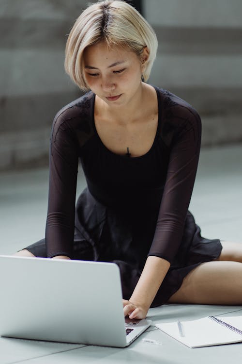 Concentrated Asian female in black ballet outfit working on netbook while sitting on floor in dance studio