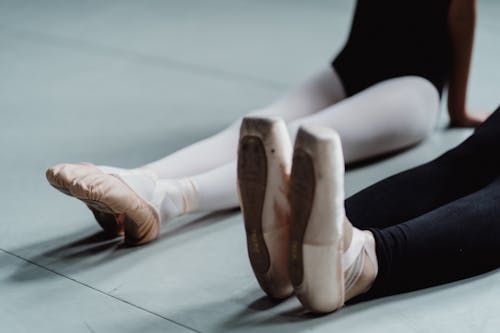 Crop unrecognizable female ballet master with little girl wearing pointes and leggings sitting on floor and doing stretching exercise during training