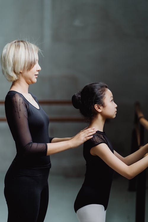 Side view of young ethnic female instructor with blond hair helping little girl doing barre exercise in studio