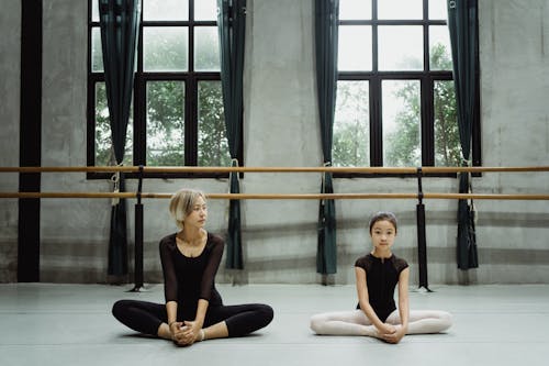 Full body of concentrated Asian little girl with female ballet teacher sitting on floor in Butterfly pose and stretching hips during training