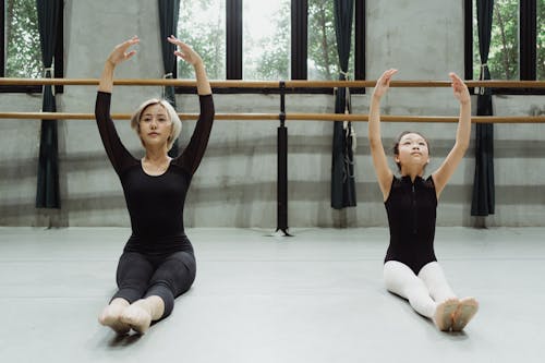 Professional Asian ballerina with beginner exercising in studio sitting on floor with raised arms