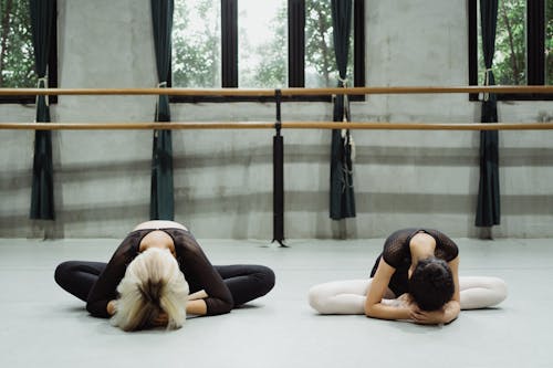 Ballet teacher with girl doing exercise with bowed heads on floor in studio with barre in daylight