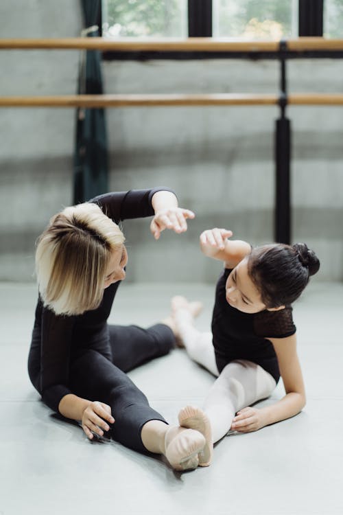 Side view of concentrated ballet teacher and focused girl stretching with raised hand together on floor in studio in daylight