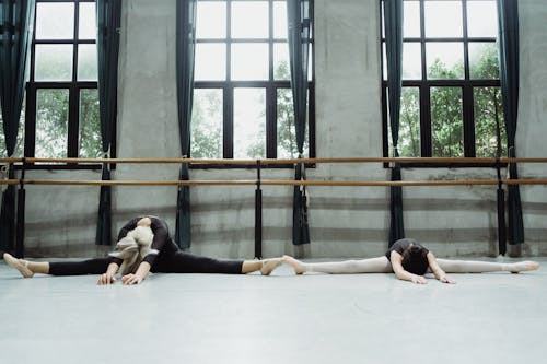 Full length anonymous woman and teenage ballerinas in leotards doing splits on floor and stretching bodies in ballet studio