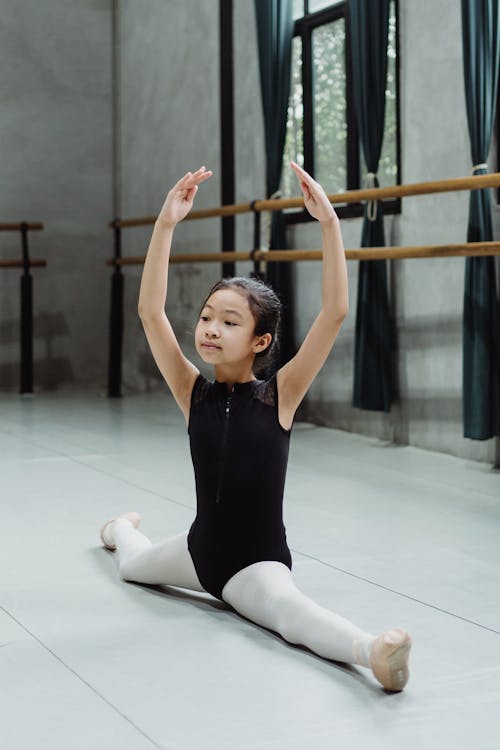 Free Full body of Asian ballet artist doing split with hands raised above head while practicing skills in classroom Stock Photo