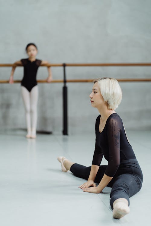 Ethnic ballerina stretching with girl in studio