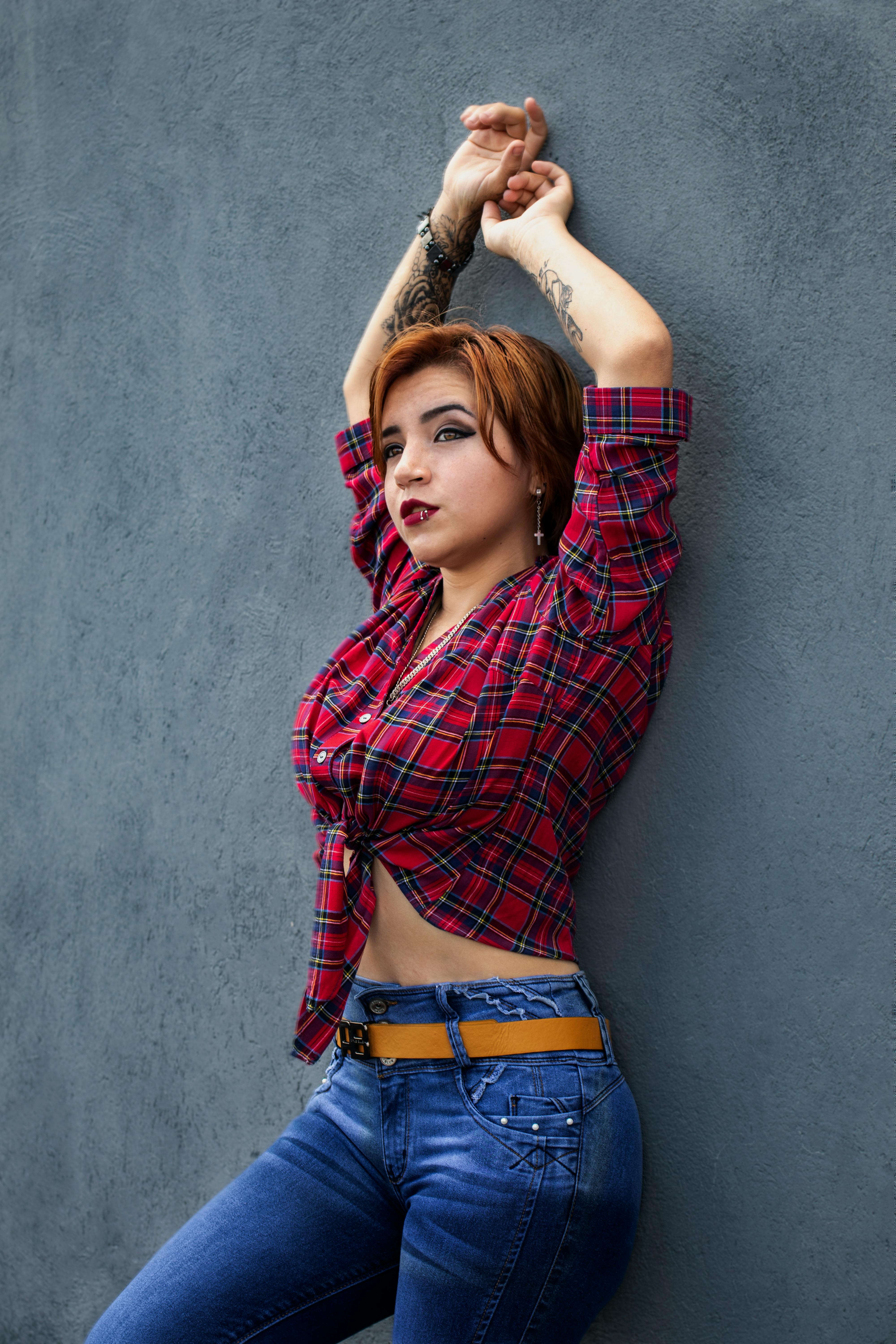 Woman in plaid shirt with tattoos raises arms against gray wall. Stylish urban portrait.