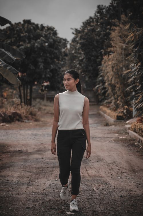 Free A young woman in casual clothing walks confidently down a serene dirt road lined with trees. Stock Photo