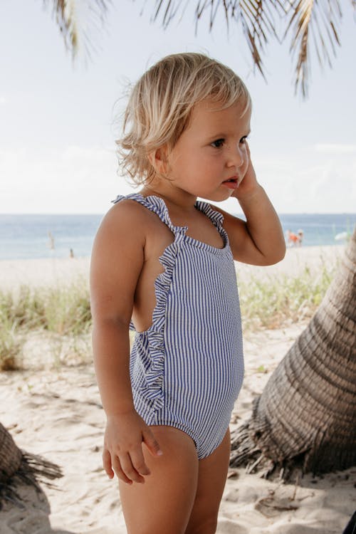 Charming little girl with short blond hair in swimsuit touching face and standing on  sandy beach near sea in sunny day