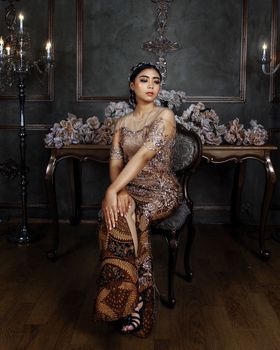 A woman in a glamorous gown sits gracefully in a richly decorated room.