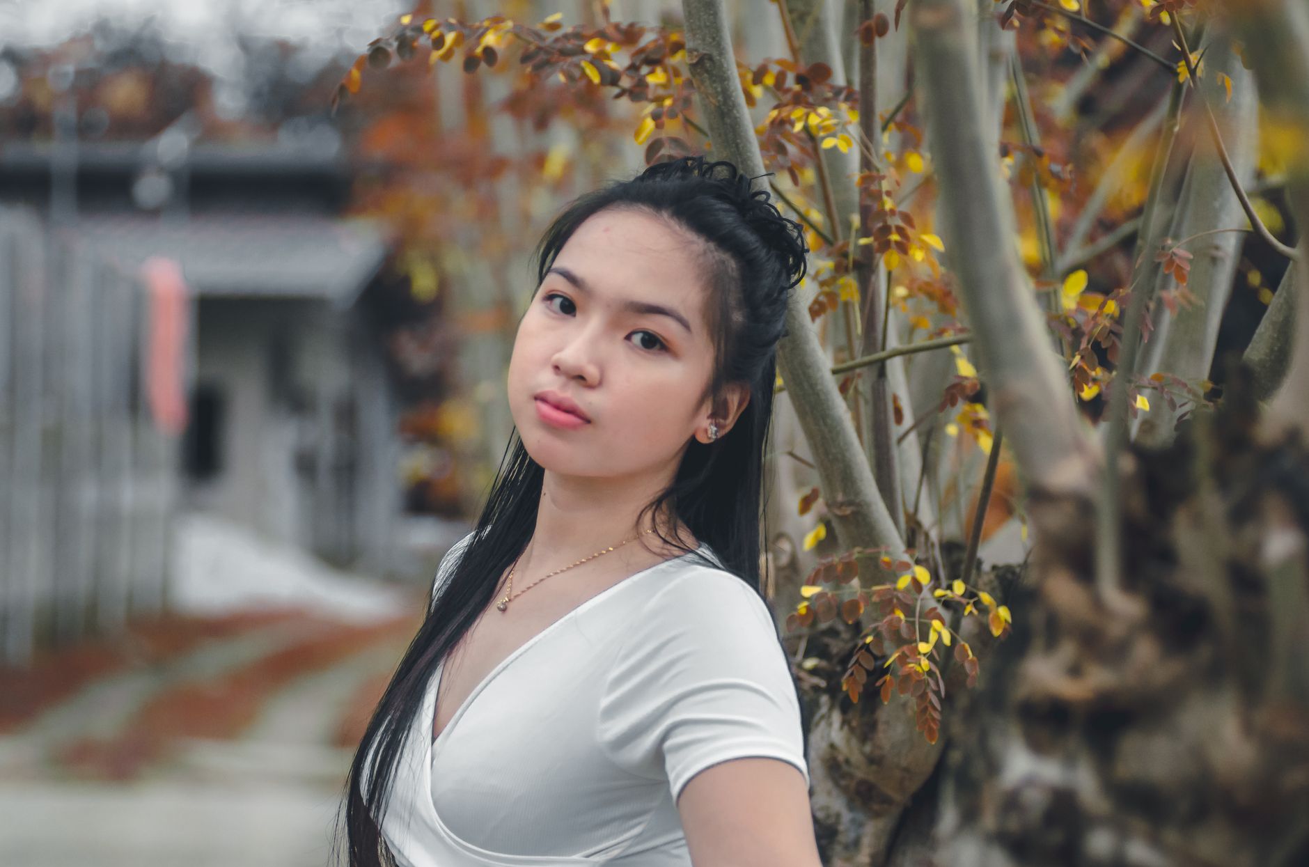 Young woman in white shirt with long hair stands outdoors in an autumn setting, surrounded by trees.
