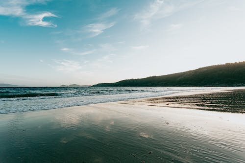 Picturesque view of seascape with rippling water washing sandy seashore near mountain ridge against cloudy sky in tropical resort in summer