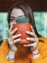 一名穿着黄色毛衣的女子在室内手捧一个装有仙人掌的陶罐，突出了她的配饰