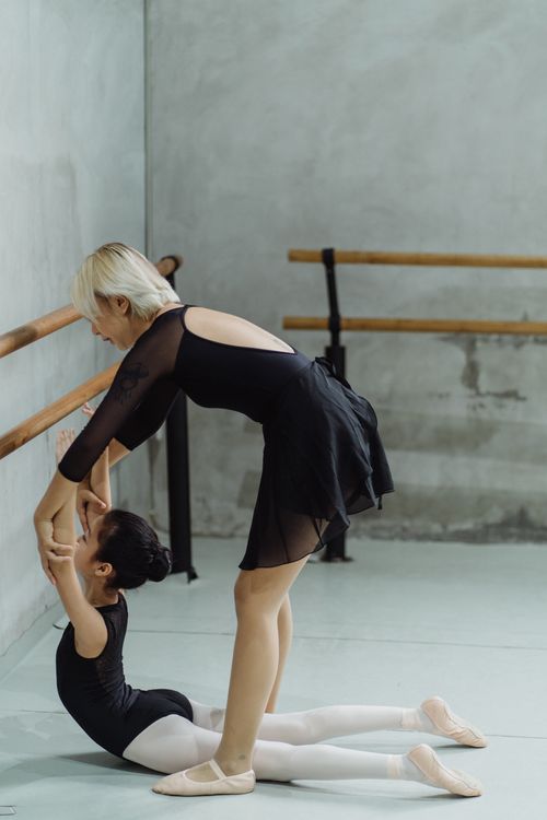 Free Side view of young unrecognizable female ballet teacher in black gymnastic costume holding arms of little ethnic girl doing back stretching exercise on floor Stock Photo