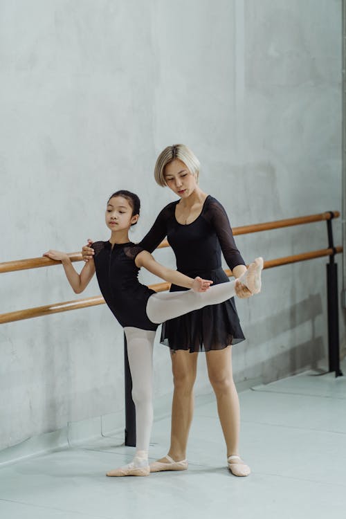 Concentrated Asian ballerina helping little girl to do choreographic movement near barre