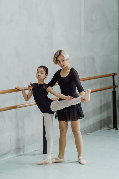 Free Full body of young ethnic female dance teacher holding leg of beginner ballerina doing stretching exercise near barre in studio Stock Photo
