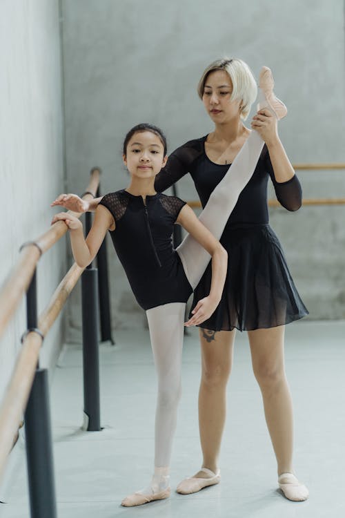 Full body of positive little ethnic girl in pointe shoes and bodysuit standing near barre and performing Developpe ballet pose with help of Asian female instructor