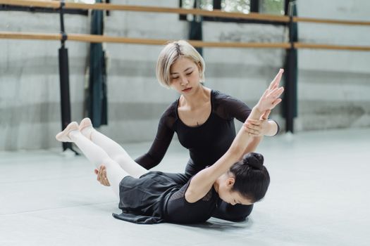 Young professional dancer helping Asian girl stretching back during ballet exercise in dancing hall