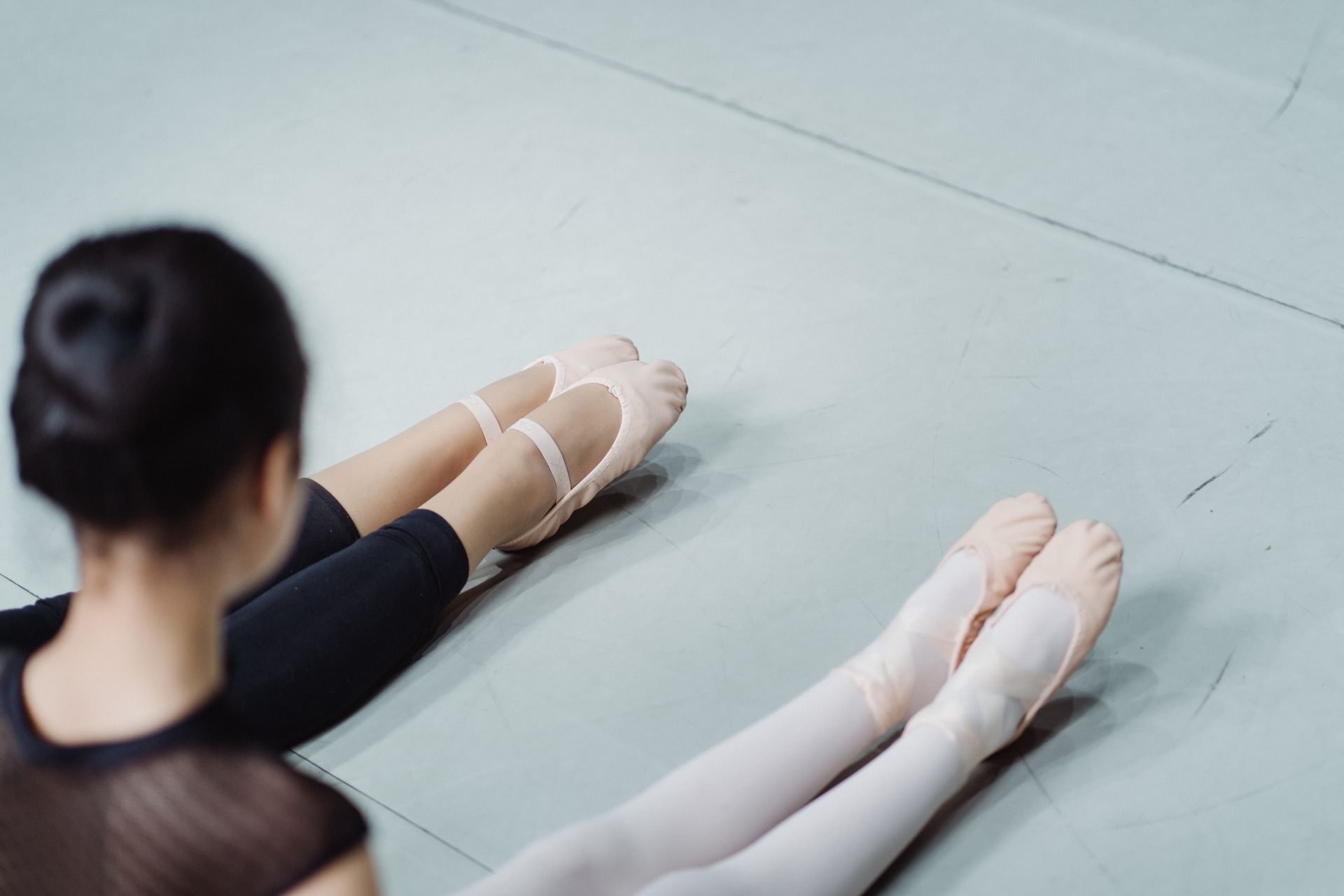 Two ballerinas stretching in a ballet class setting, focusing on their legs in pointe shoes.
