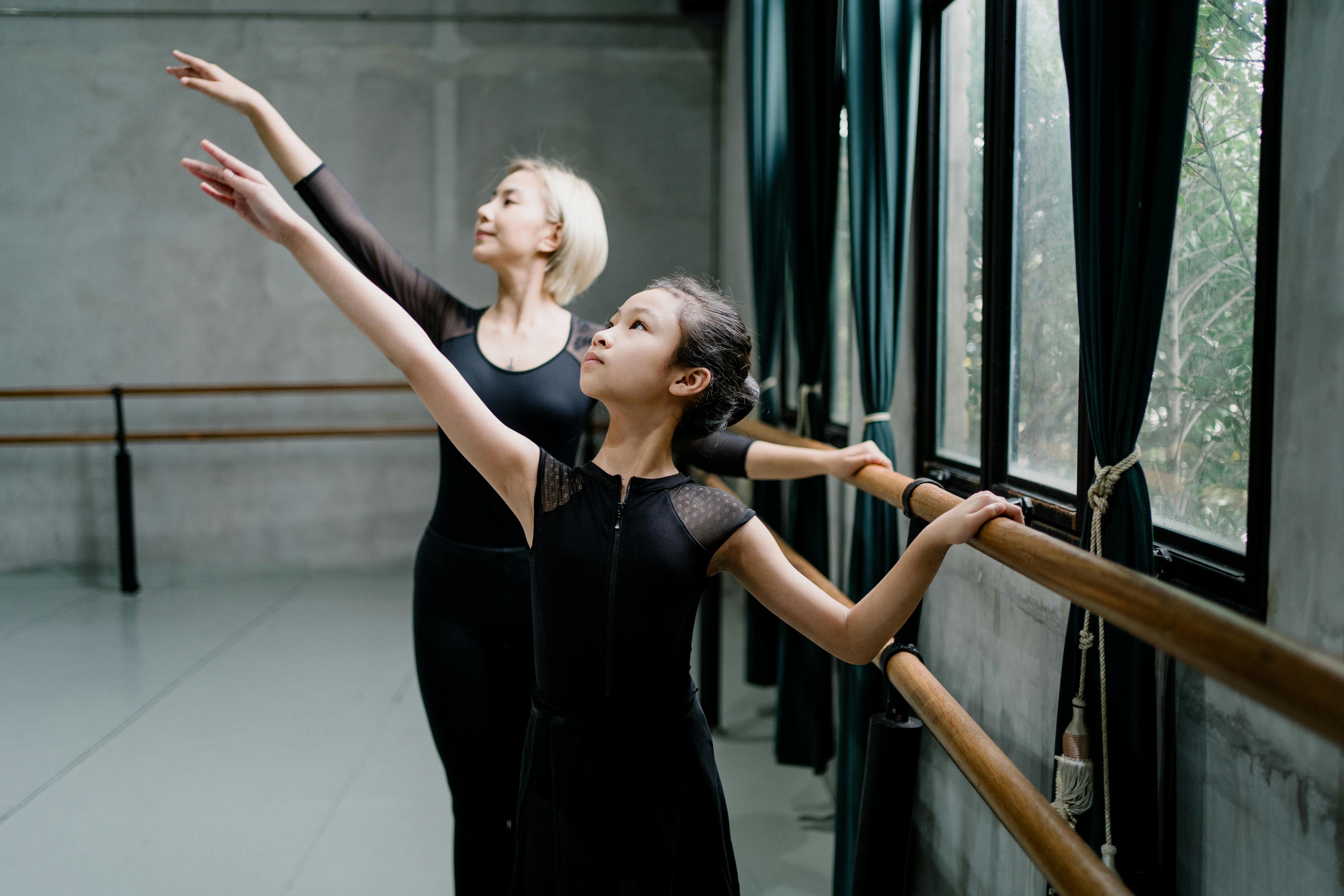 positive asian ballerinas standing near barre and raising arms gracefully