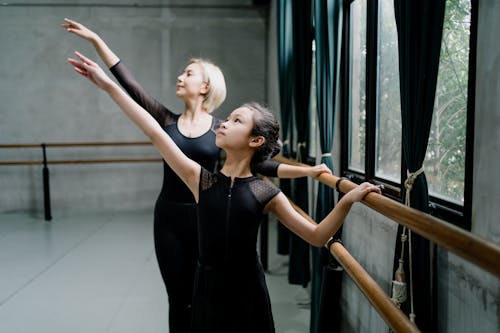 Slender Asian girl ballerina and female ballet instructor in black leotards practicing near barre and raising arms gracefully during rehearsal in modern ballet studio