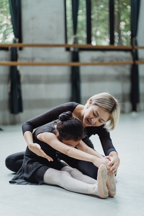 Full length professional Asian female ballet instructor in leotard sitting on floor and helping girl ballerina to stretch body in light studio