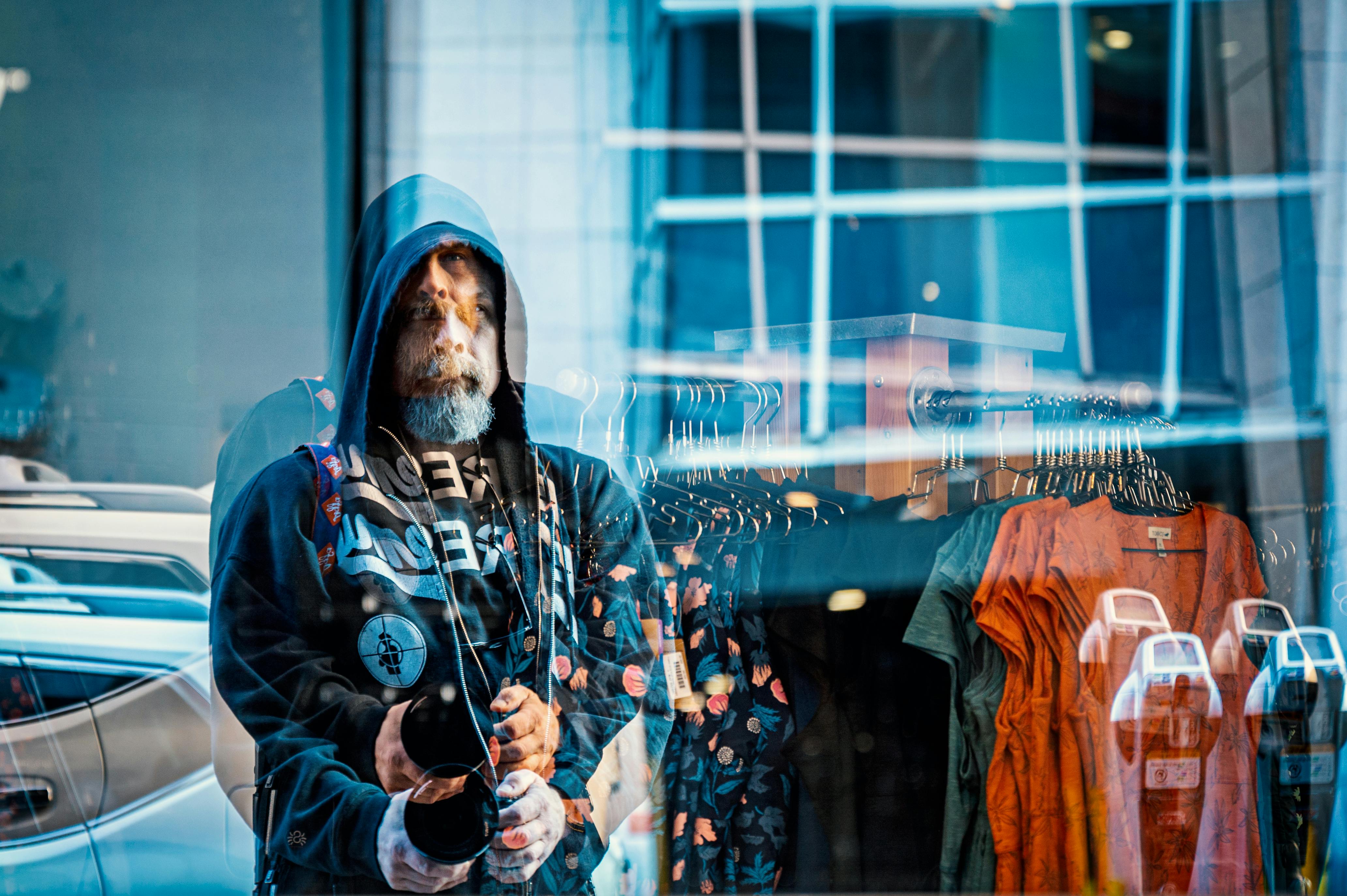Through glass of middle aged serious bearded brutal male in hoodie customer choosing clothes hanging on rack in modern boutique