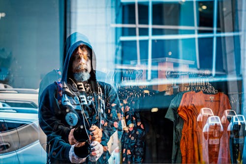 Through glass of middle aged serious bearded brutal male in hoodie customer choosing clothes hanging on rack in modern boutique