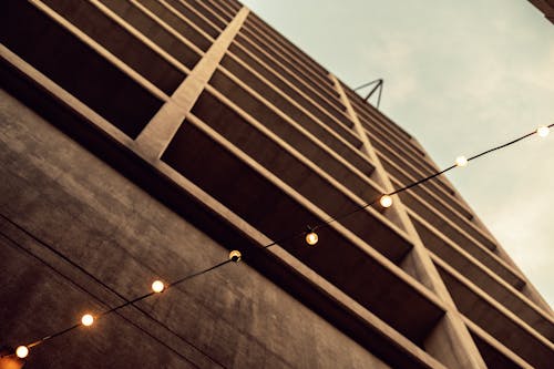 Low angle of cable with glowing light bulbs hanging on street near multistory residential building with balconies against cloudy sky