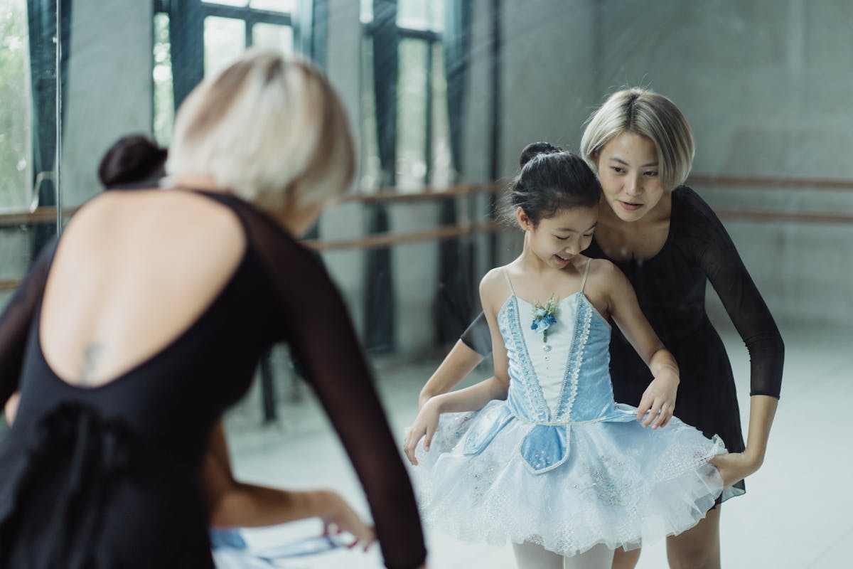 Positive Asian female ballet instructor standing with girl trainee against mirror and adjusting white tutu skirt