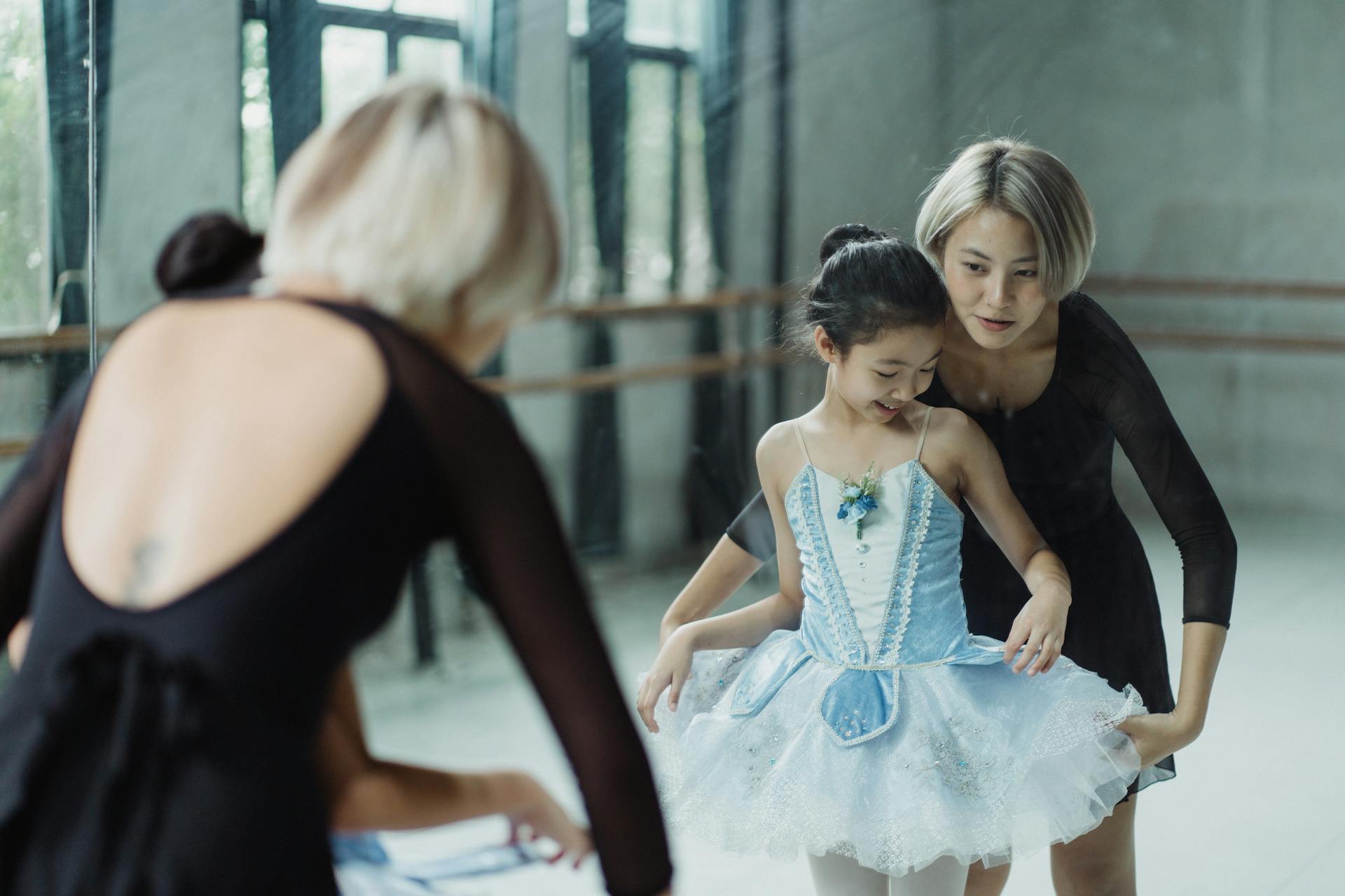 Positive Asian female ballet instructor standing with girl trainee against mirror and adjusting white tutu skirt