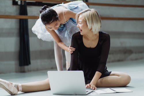 Joyful Asian female ballet instructor and girl ballerina in tutu browsing netbook on floor in light modern studio