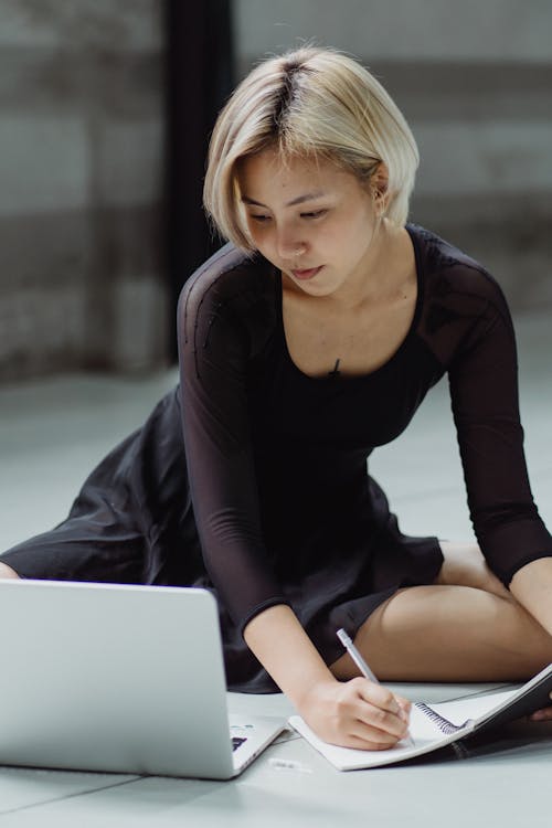 Focused young Asian female in black dress sitting on floor and taking notes in copybook while browsing netbook