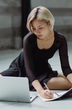 一位身着黑色礼服的年轻亚洲女性坐在地板上，一边浏览上网本，一边在字帖上做笔记