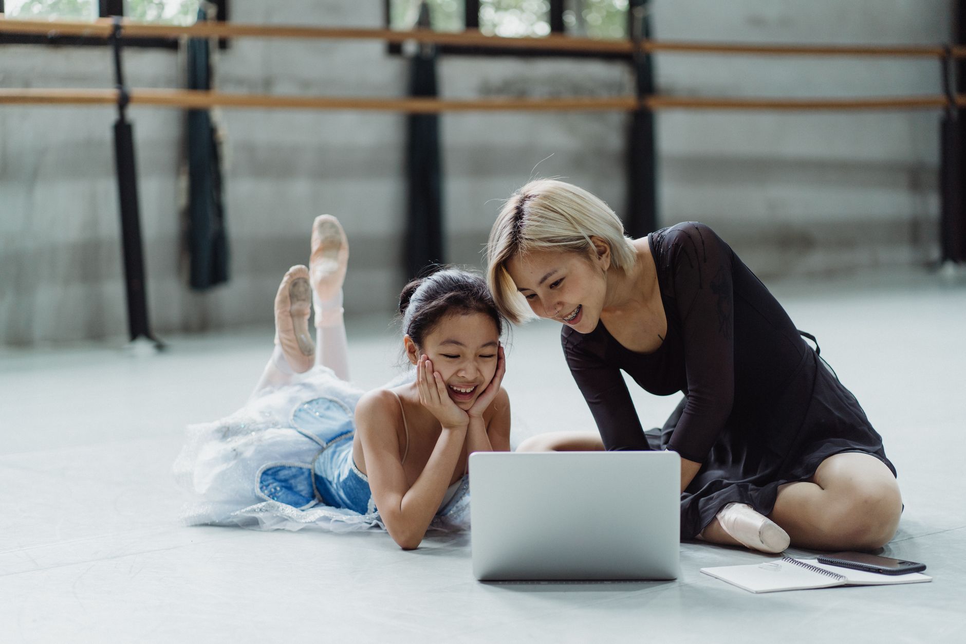 Full body of positive Asian female ballet instructor watching netbook sitting near girl in tutu skirt