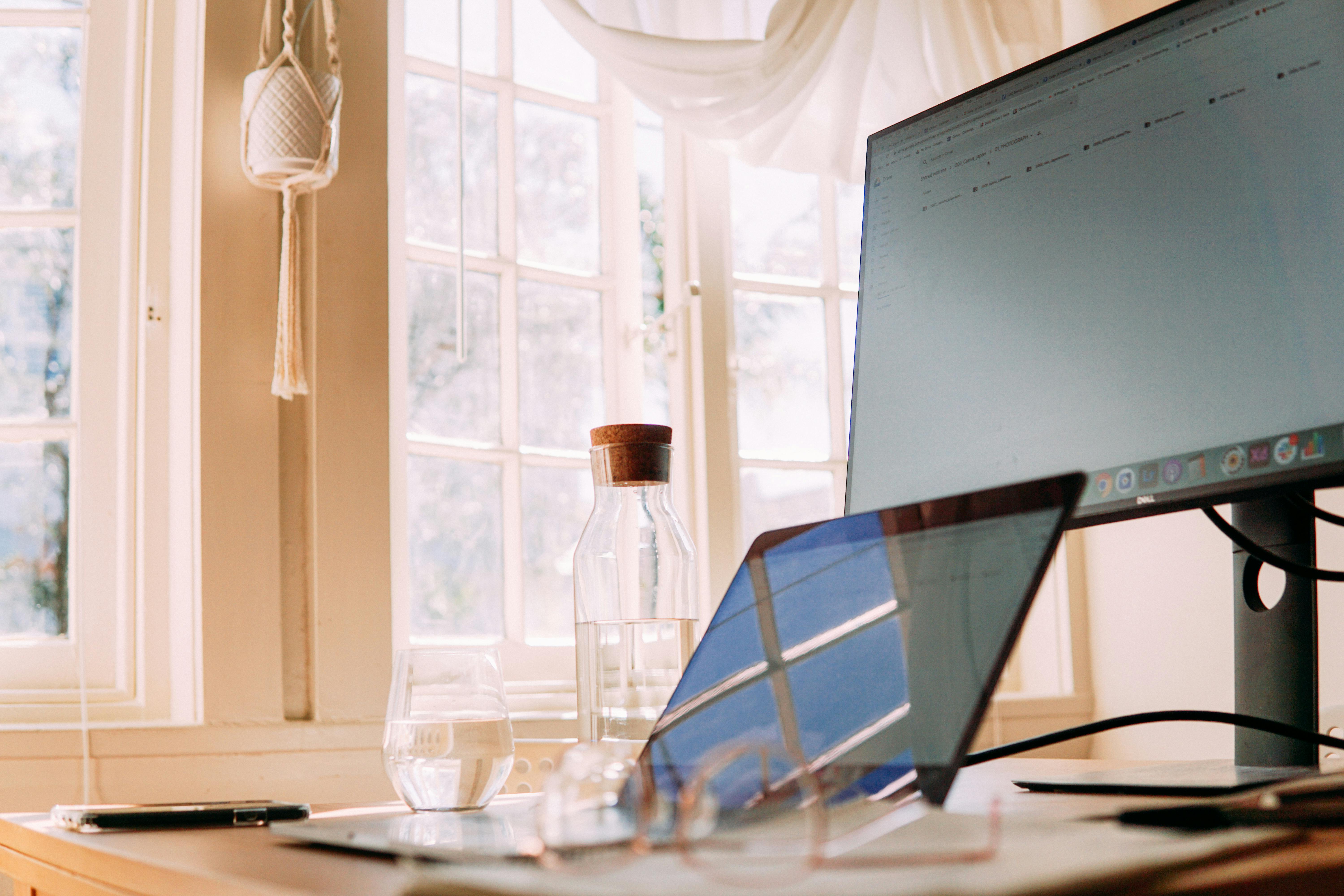 A modern home office setup with a laptop and monitor, bathed in natural light through large windows.