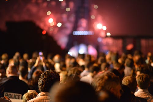 This photo displays a crowd of people who stand in front of the Eiffel Tower and who are watching the red colors of fireworks which seems to be happening behind and around the Eiffel Tower. On the lower left side a policeman is visible.