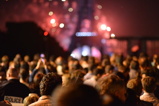 This photo displays a crowd of people who stand in front of the Eiffel Tower and who are watching the red colors of fireworks which seems to be happening behind and around the Eiffel Tower. On the lower left side a policeman is visible.