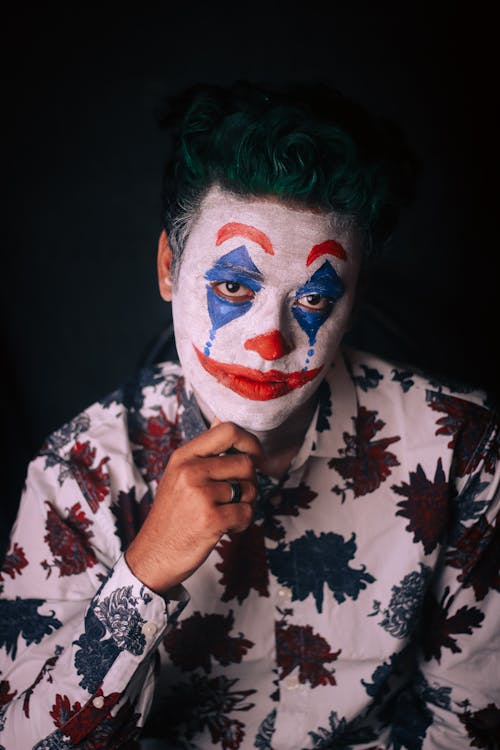 Pensive male in shirt with floral print with green hair and greasepaint on face touching chin and looking at camera on dark background