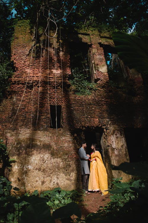 Full body of Indian couple in festive suits looking at each other while standing in doorway old abandoned construction with destructed shabby brick walls under foliage of tall green trees