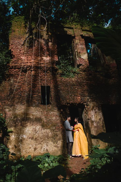 Free Full body of Indian couple in festive suits looking at each other while standing in doorway old abandoned construction with destructed shabby brick walls under foliage of tall green trees Stock Photo