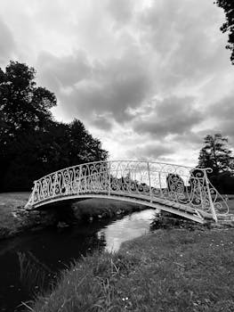 A classic black and white photo of an elegant iron bridge over a tranquil pond in a serene park.