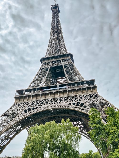 Free View of the Eiffel Tower against a cloudy sky, framed by greenery at the base. Stock Photo